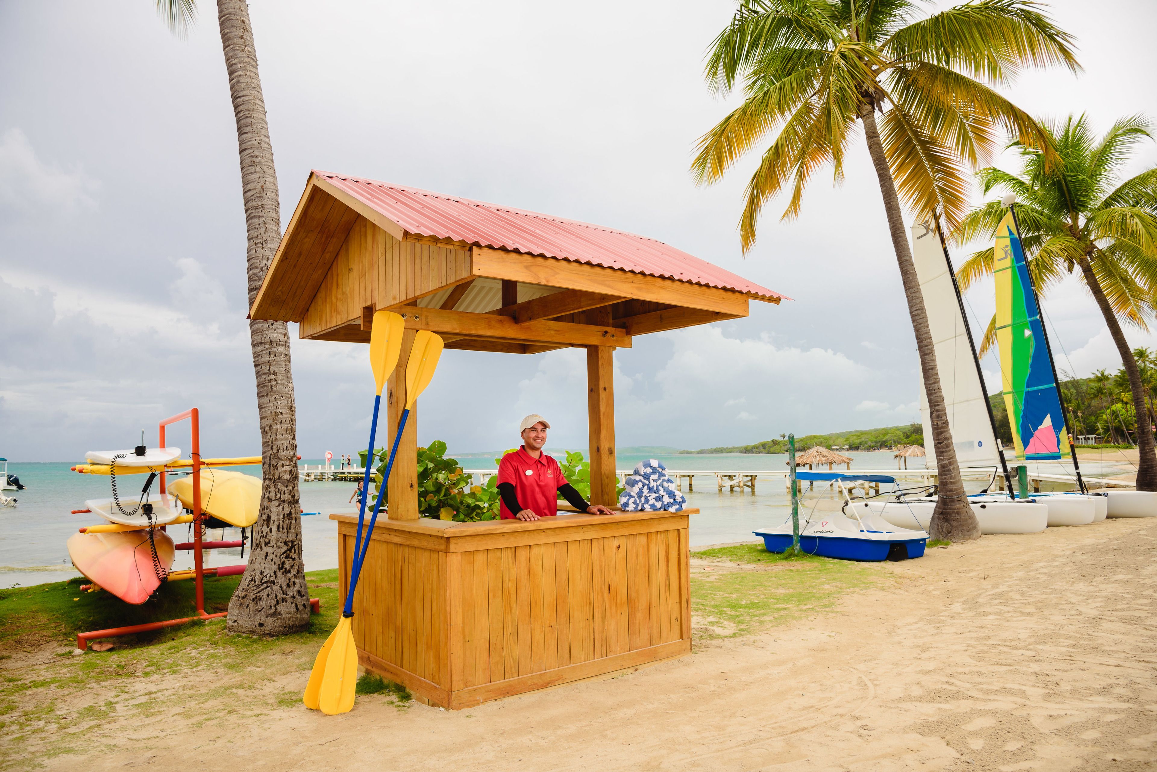 A wooden beach kiosk with a red roof, surrounded by palm trees and a beach view.