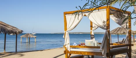 A wooden bed with a canopy, a table with bottles, and a thatched umbrella on a sandy beach with the sea in the background.