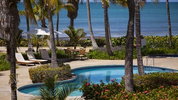 A pool area with lounge chairs and umbrellas, surrounded by palm trees and a view of the ocean.