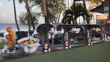 Un bufé con diversos alimentos y condimentos, con vista a una playa con palmeras.