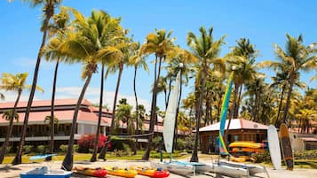 Una playa con palmeras, un edificio de techo rojo y varios kayaks y un velero.