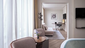 Ein modernes Hotelzimmer mit einer Couch, einem kleinen runden Tisch mit Blumenvase, einem Schreibtisch mit Lampe und einem flachbildfernseher an der Wand.