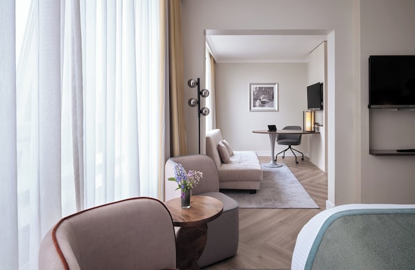 Ett modernt hotellrum med en soffa, ett litet runt bord med en vas med blommor, ett skrivbord med en lampa och en platt-TV som är monterad på väggen.