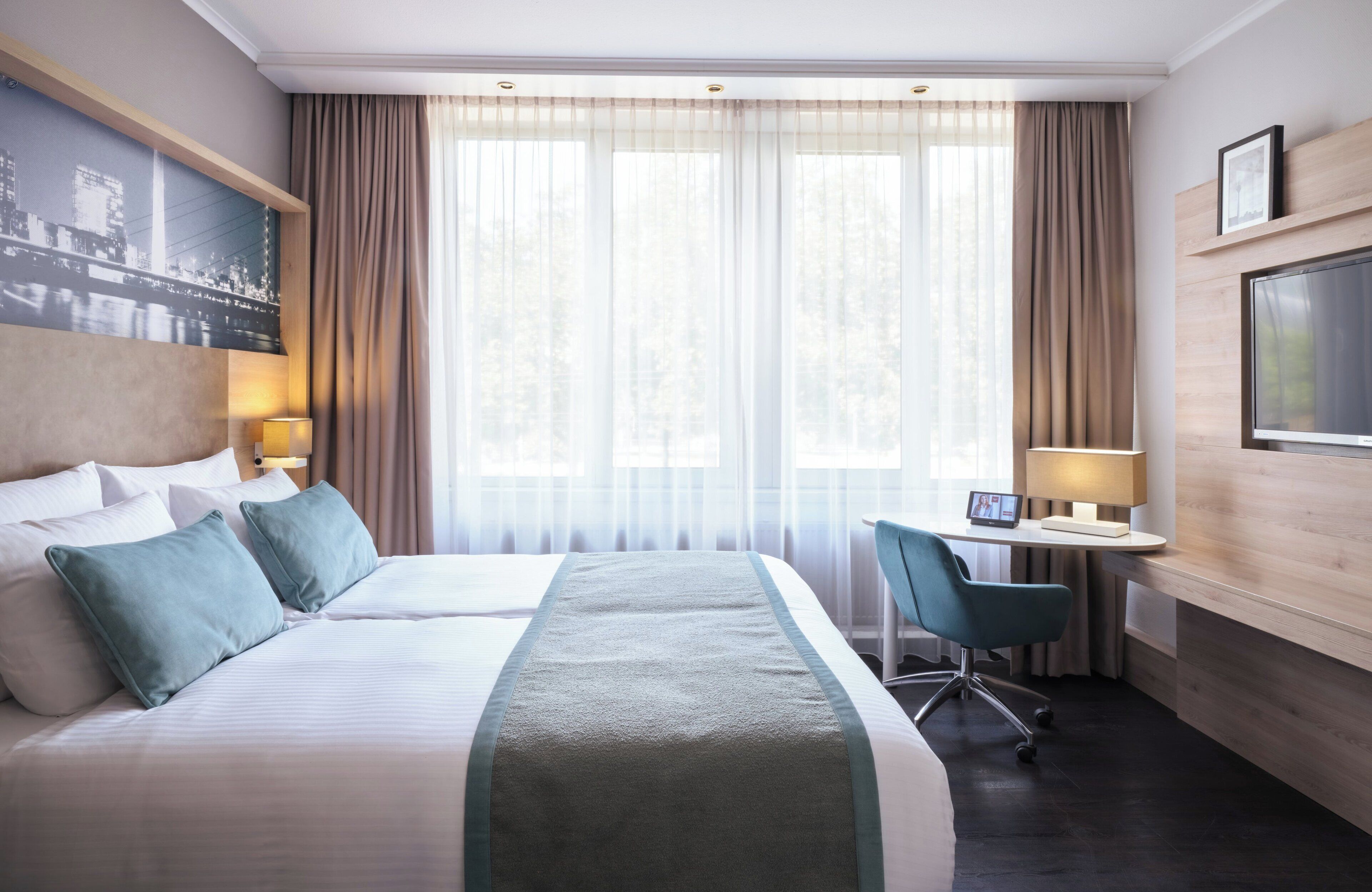 Quarto de hotel com uma cama grande, uma escrivaninha com cadeira, uma TV e uma pintura de paisagem urbana na parede.