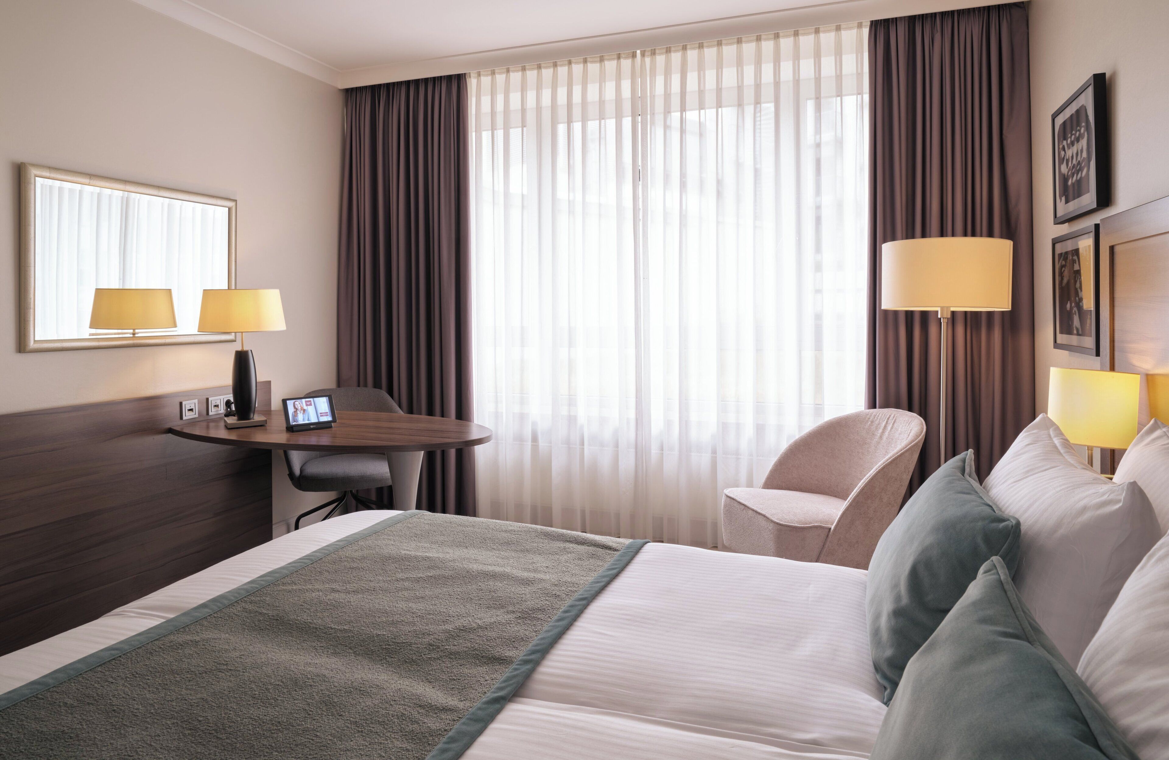 A hotel room with a large bed, a desk with a computer, a chair, and a window with curtains.