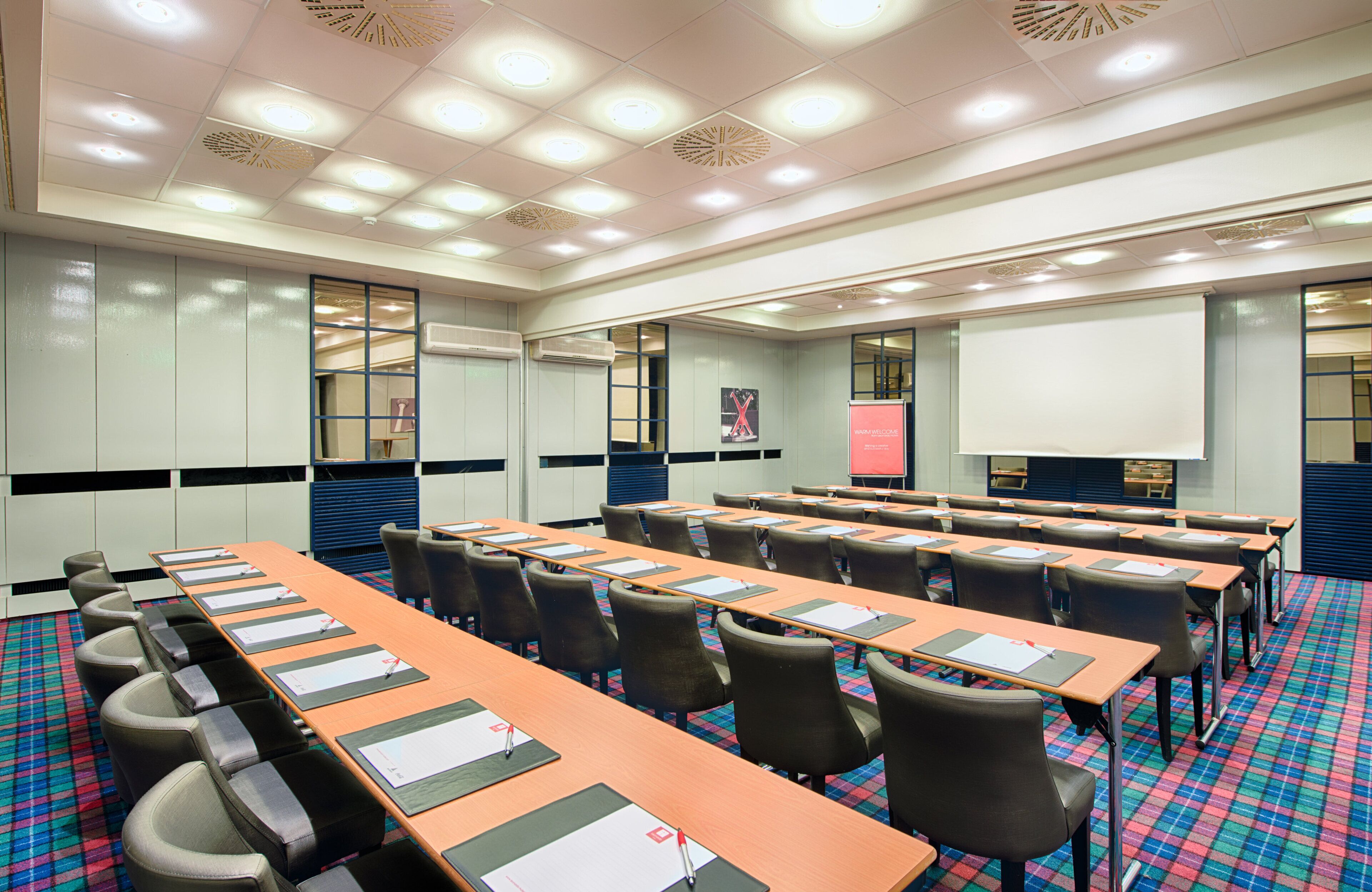 A conference room with a long table, chairs, a projector screen, and a 'Warm Welcome' sign.