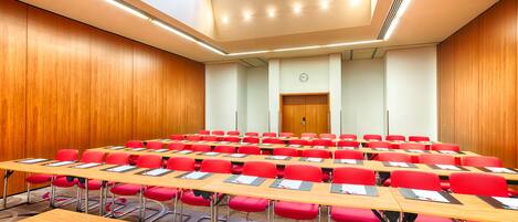 A conference room with rows of red chairs and wooden tables, equipped with nameplates and pens.