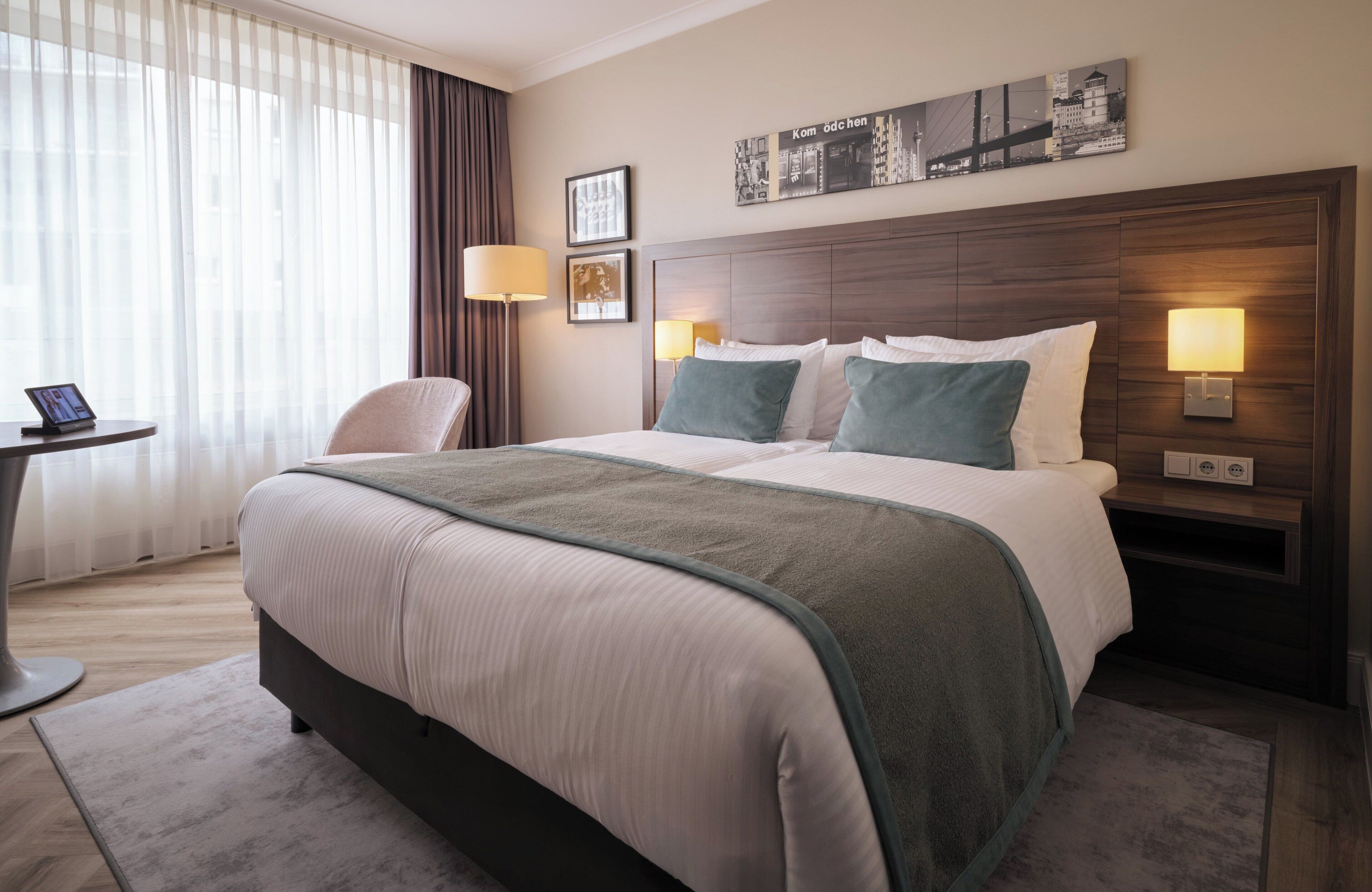 A hotel room with a large bed, a desk with a lamp, and a view of the city through the window.