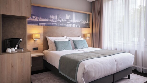 Ein modernes Hotelzimmer mit einem großen Bett, zwei Nachttischen mit Lampen, einem Holzschrank und einem Wandbild mit Stadtansicht.