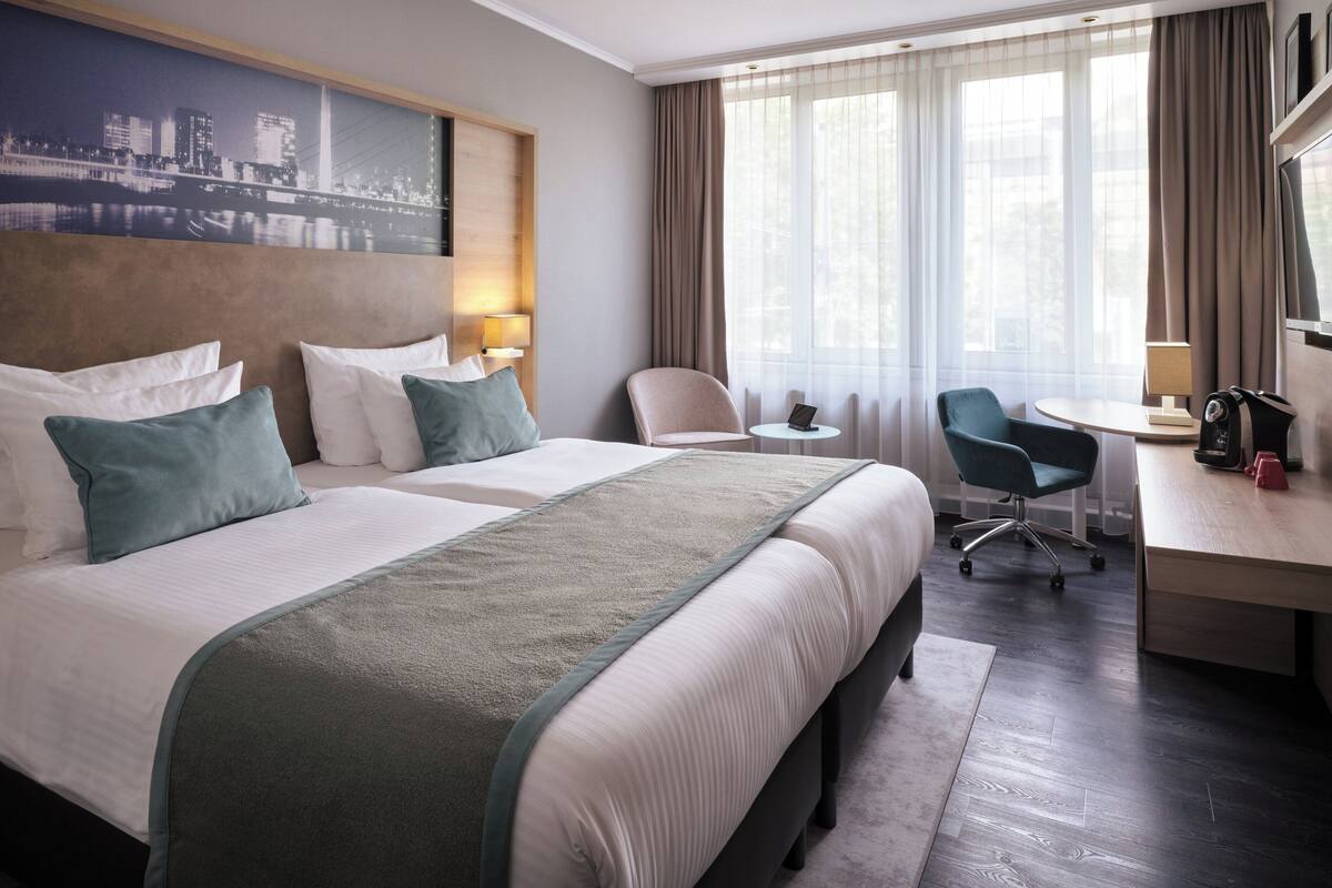 Habitación de hotel con dos camas, un escritorio, una silla, una mesita y vista a la ciudad.