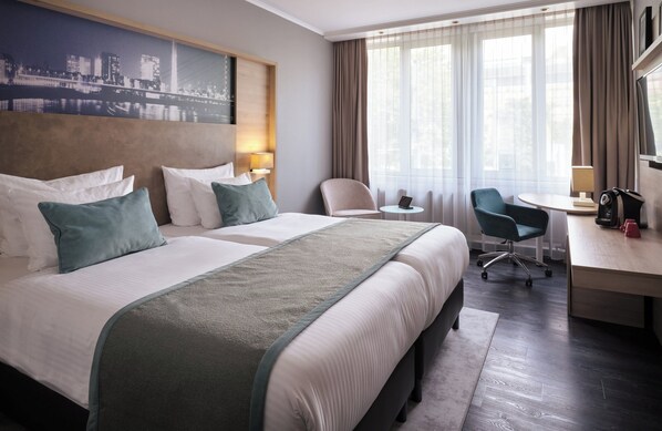 A hotel room with two beds, a desk, a chair, a small table, and a cityscape view. - Leonardo Royal Hotel Düsseldorf Königsallee (Düsseldorf)
