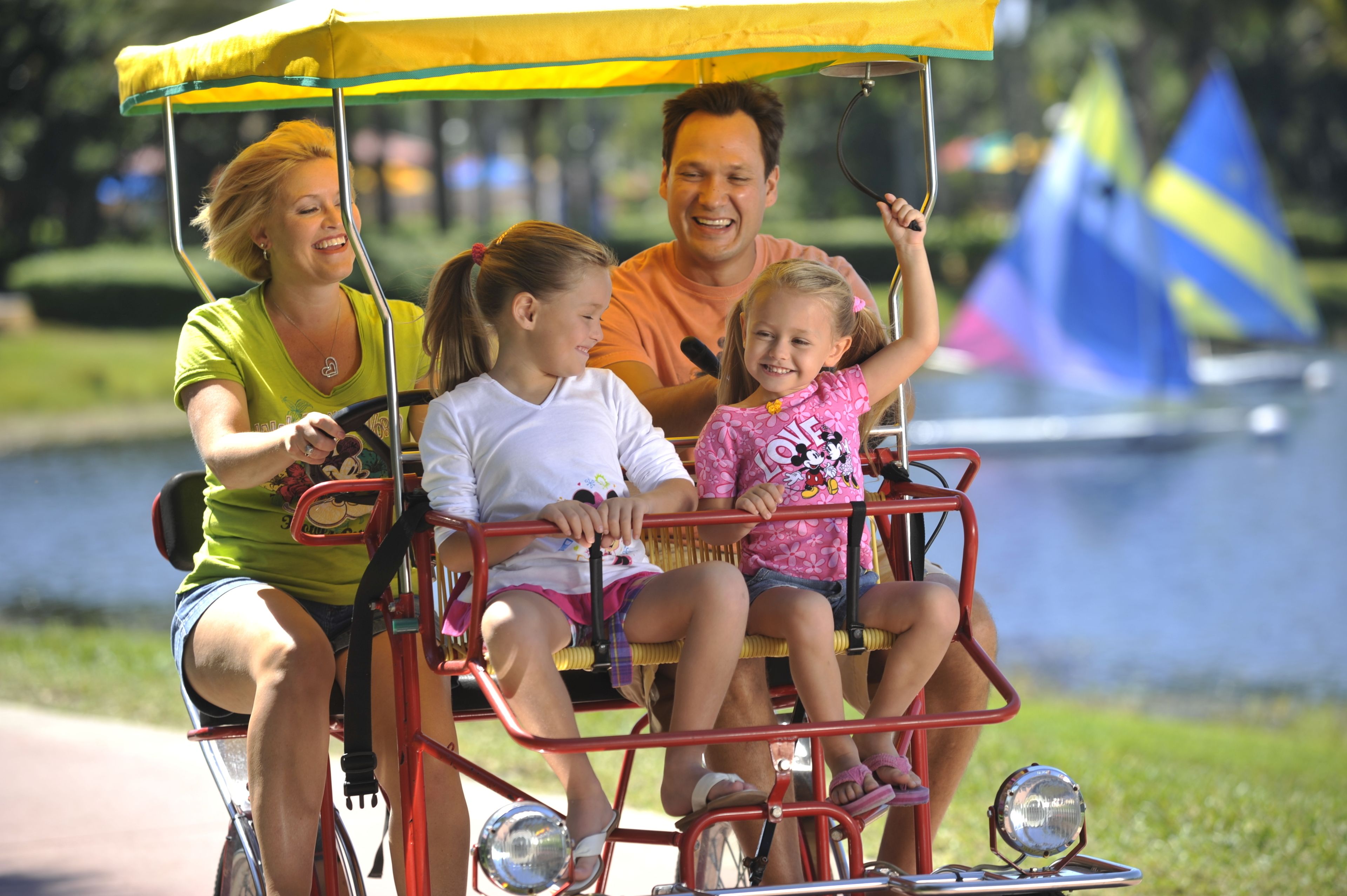 A family on a red and yellow trolley with a canopy, riding along a path near a body of water.
