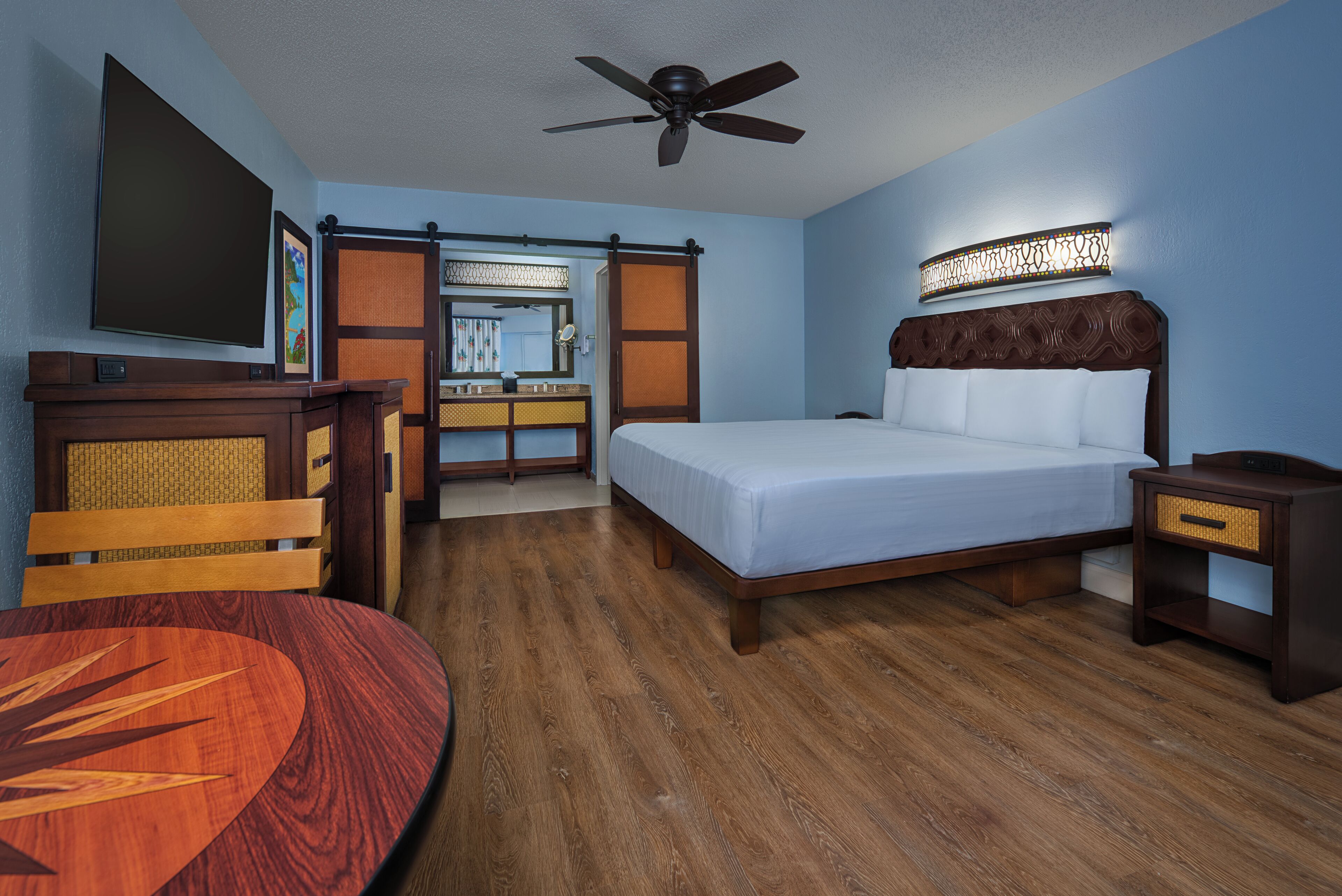 A hotel room with a bed, a desk with a chair, a dining table, a ceiling fan, and a bathroom with a mirror and sink.