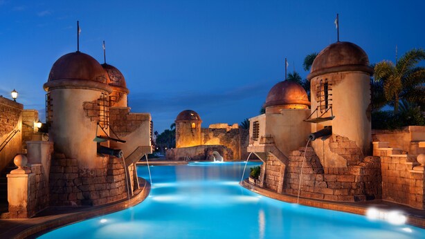A pool area with stone structures and domed towers, surrounded by palm trees. - Disney's Caribbean Beach Resort (Lake Buena Vista)
