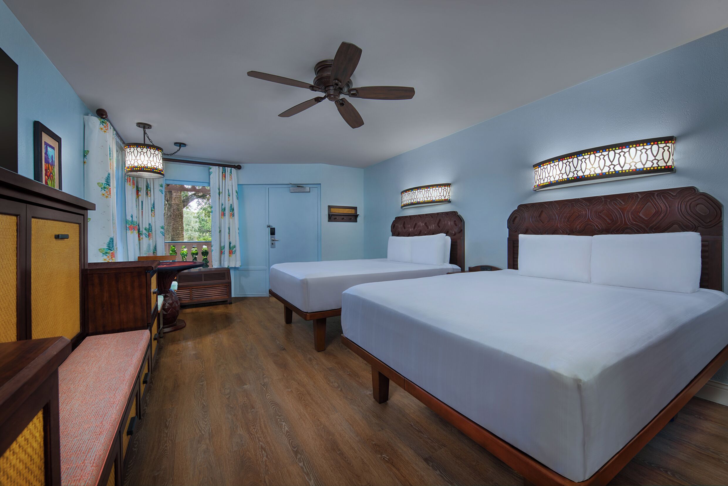 A hotel room with two beds, a ceiling fan, a wooden bench, a desk, and a window with curtains.