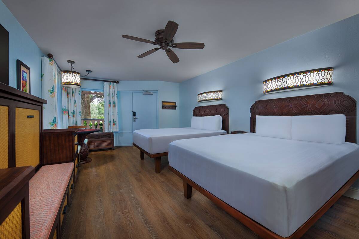 A hotel room with two beds, a ceiling fan, a wooden bench, a desk, and a window with curtains.