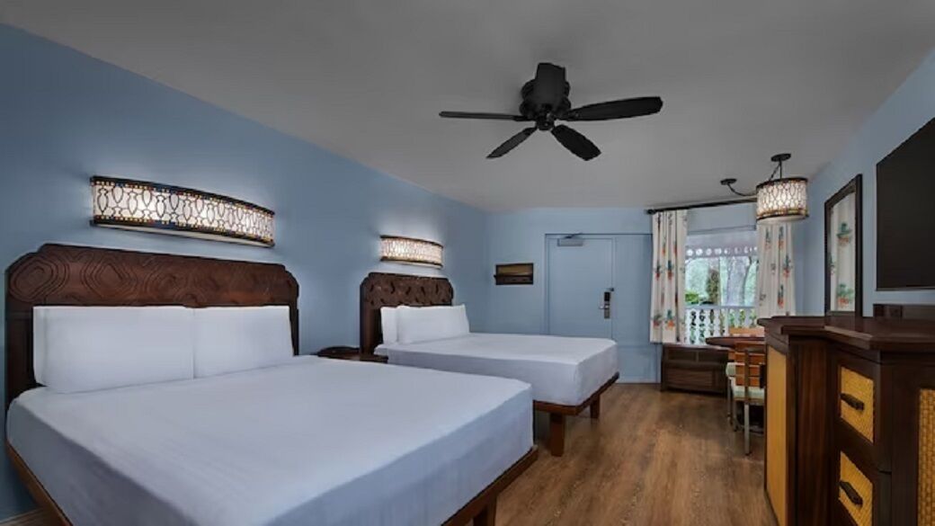 A hotel room with two beds, a ceiling fan, a desk with a chair, and a TV.
