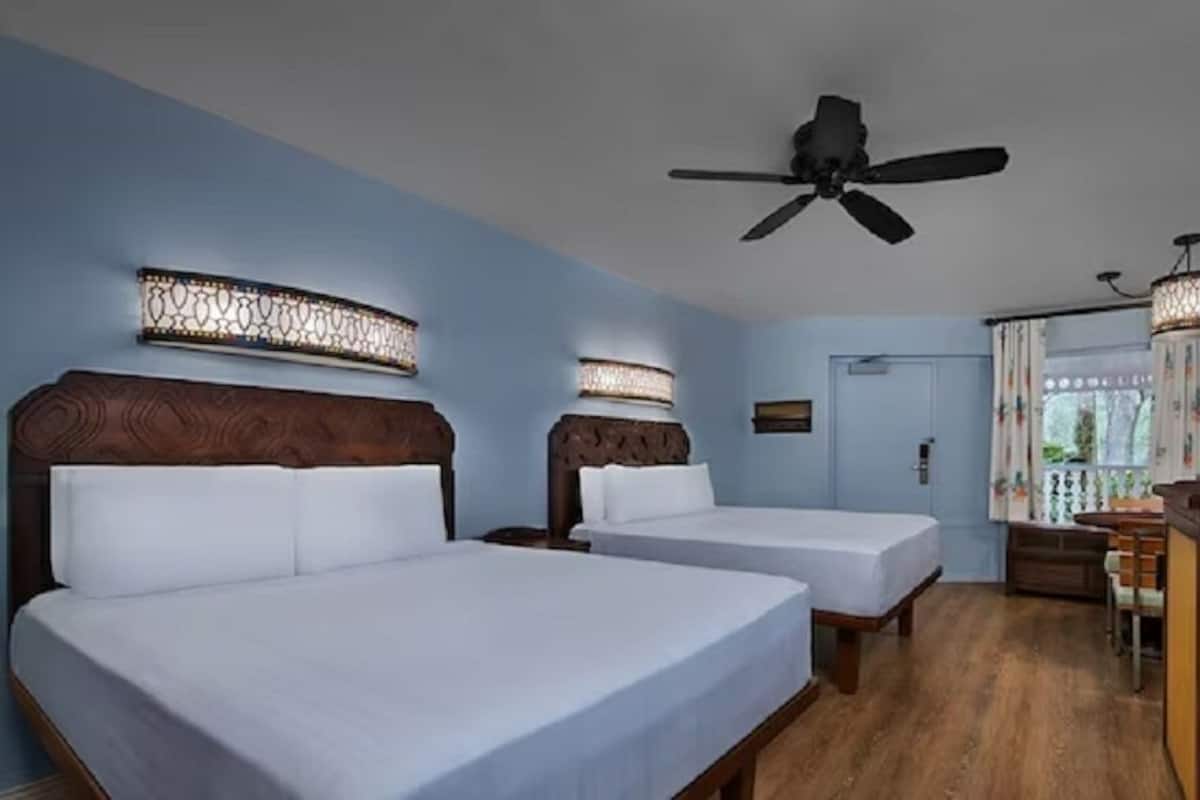 Habitación de hotel con dos camas, ventilador de techo, escritorio con silla y televisor.