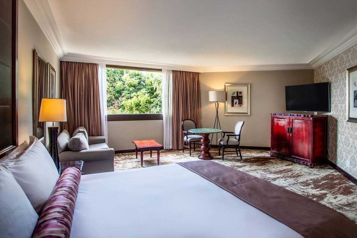 Une chambre d’hôtel avec un grand lit, un bureau, une chaise, une télévision et une fenêtre avec des rideaux.