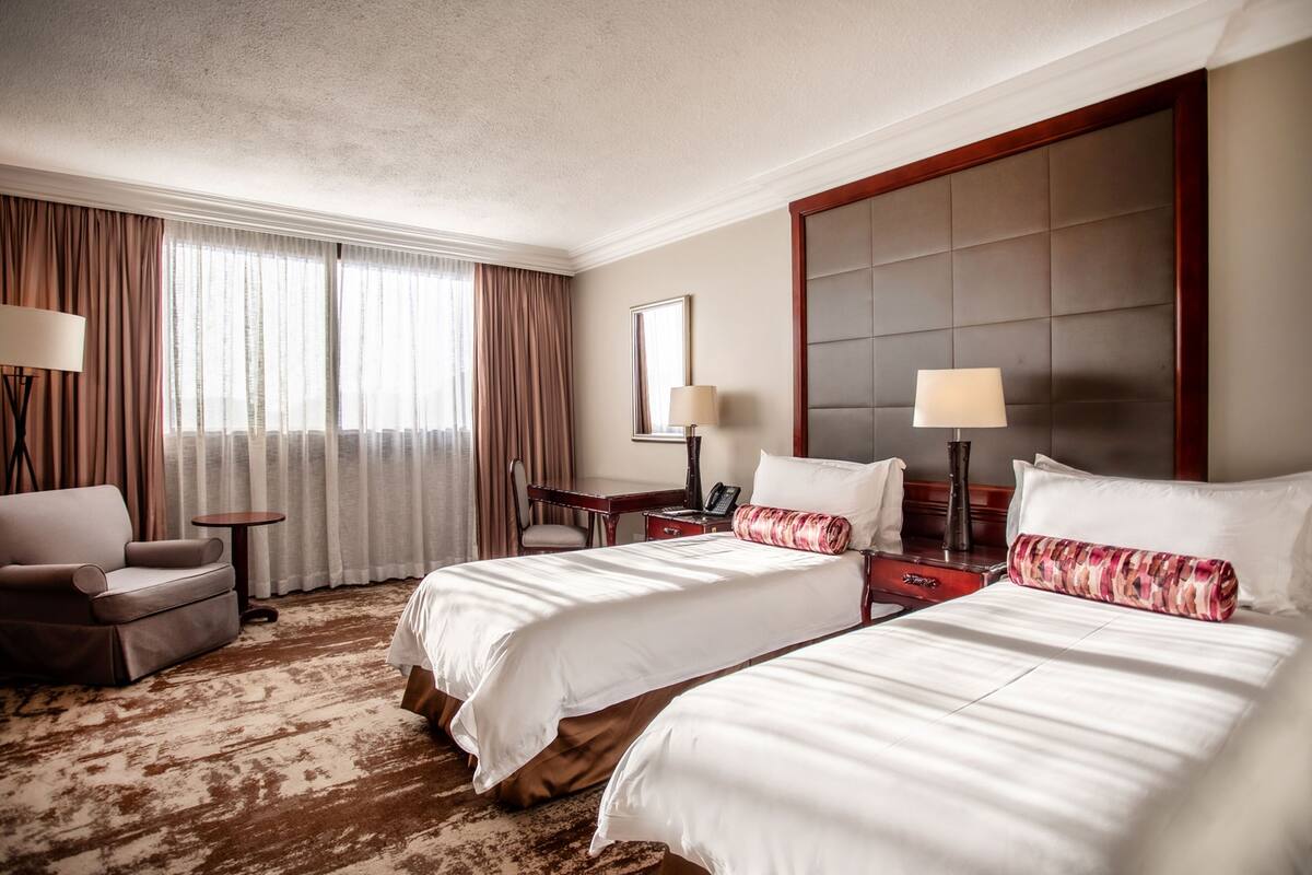 Une chambre d’hôtel avec deux lits, un bureau, une chaise et une grande fenêtre avec des rideaux.
