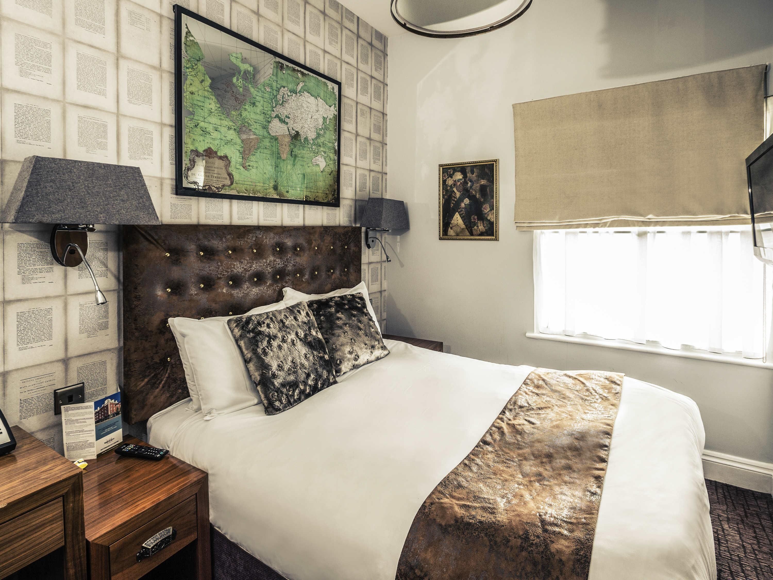 Habitación de hotel ordenada con una cama grande, un mapa en la pared, una ventana con cortinas y una mesita de noche con lámpara.