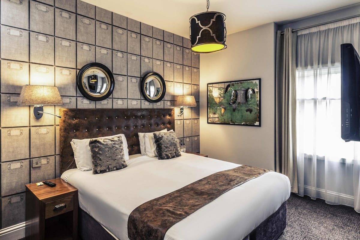 A hotel room with a large bed, two bedside lamps, a headboard with a mirror, a framed artwork on the wall, and a window with curtains.
