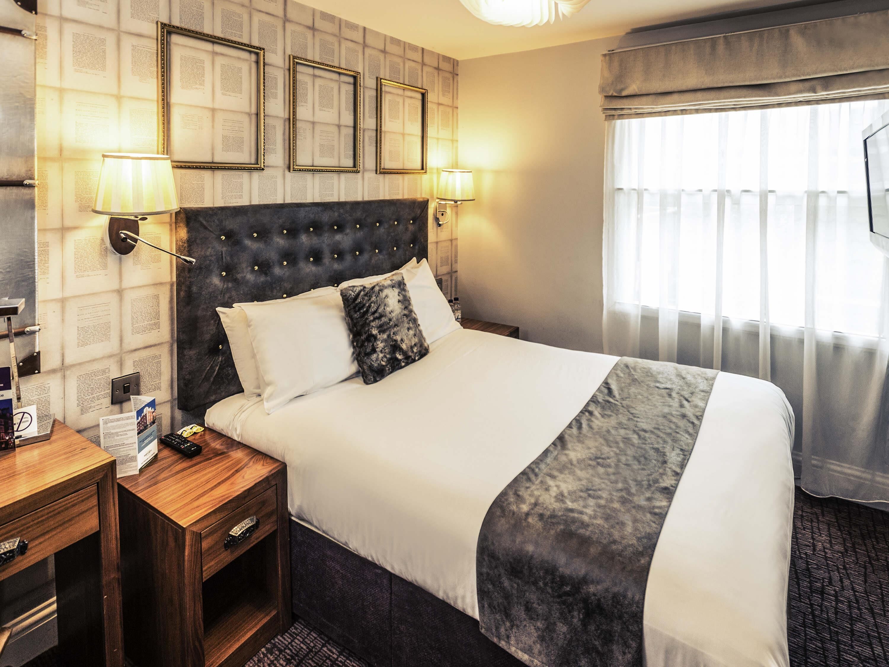 A hotel room with a large bed, two bedside lamps, a wooden nightstand, and a window with sheer curtains.