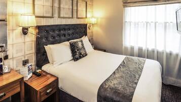 Una habitación de hotel con una cama grande, dos lámparas de noche, una mesita de noche de madera y una ventana con cortinas translúcidas.
