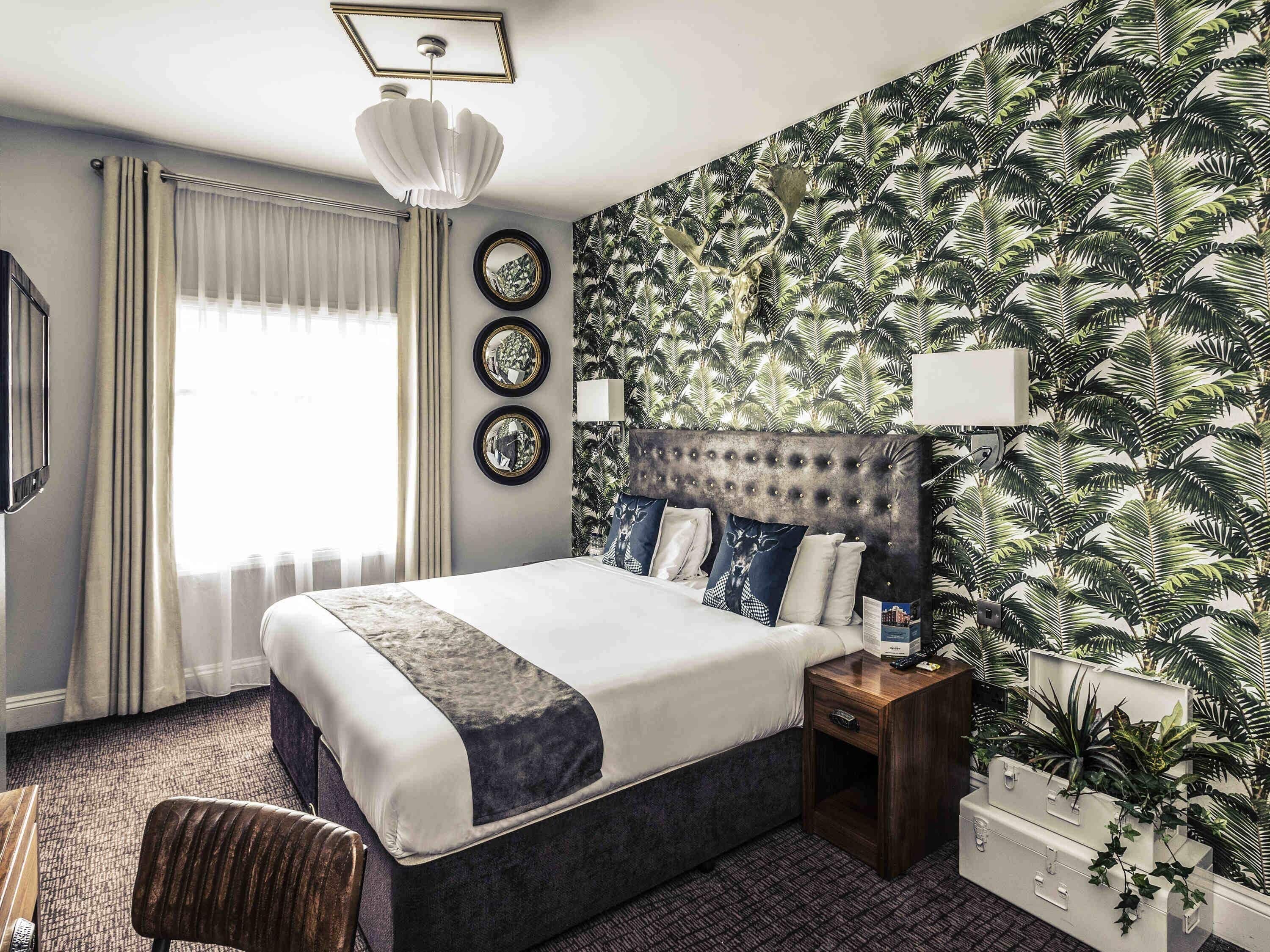 Ein Hotelzimmer mit einem großen Bett, einem Nachttisch, einem Spiegel und einer Wand mit tropischem Blattmuster.