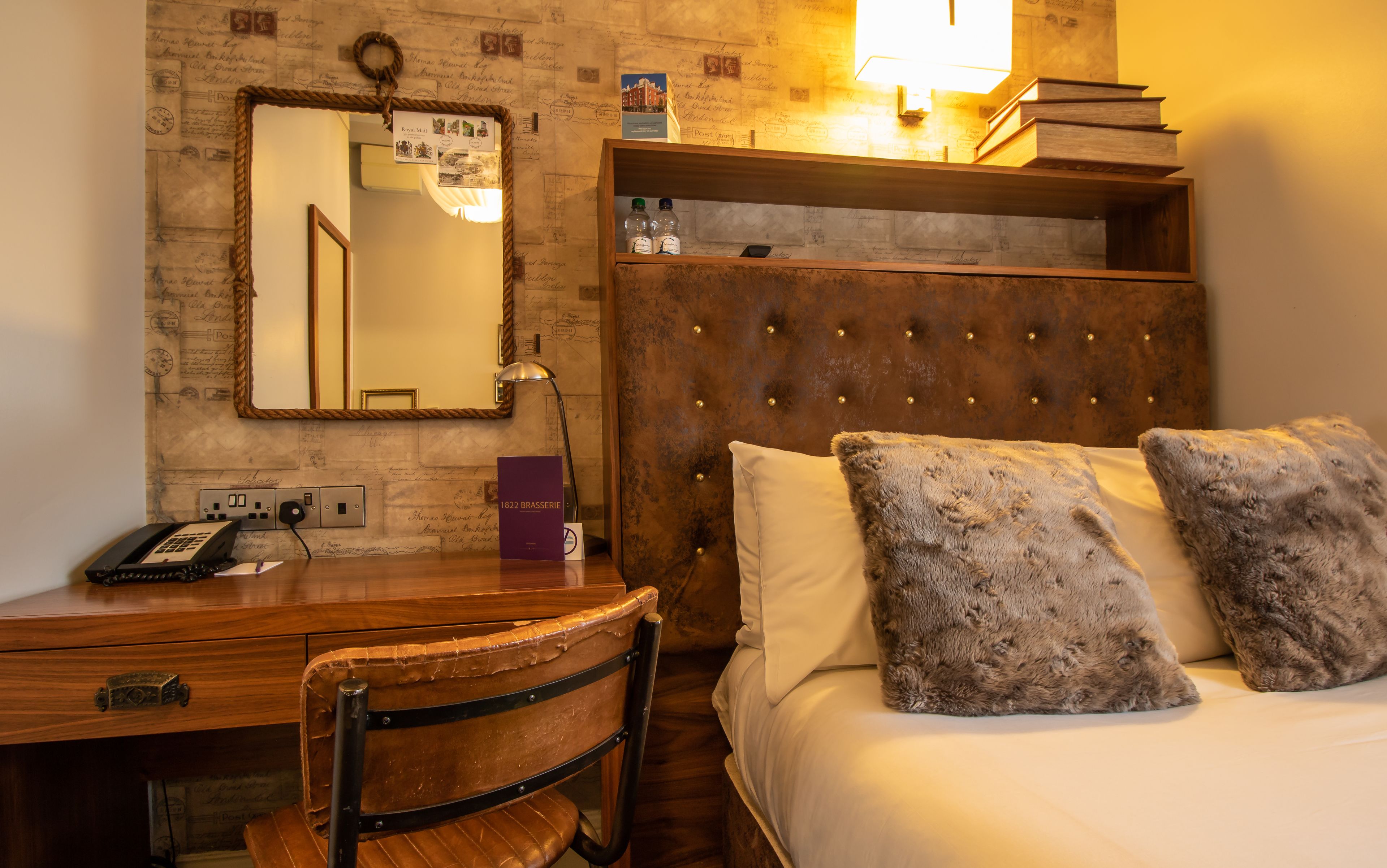 A hotel room with a wooden desk, a telephone, a mirror, a headboard with tufting, and two fluffy pillows.