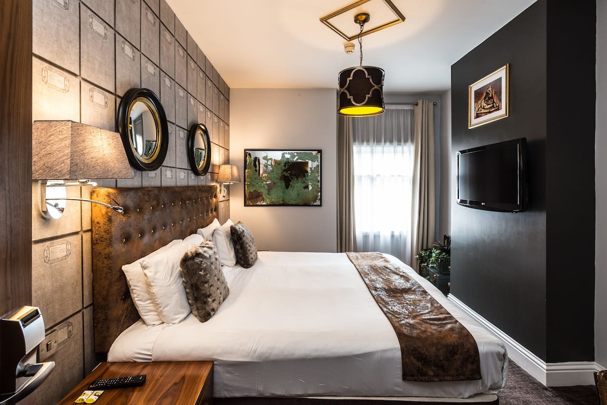 A hotel room with a large bed, two wall-mounted mirrors, a flat-screen TV, a painting on the wall, and a window with curtains.
