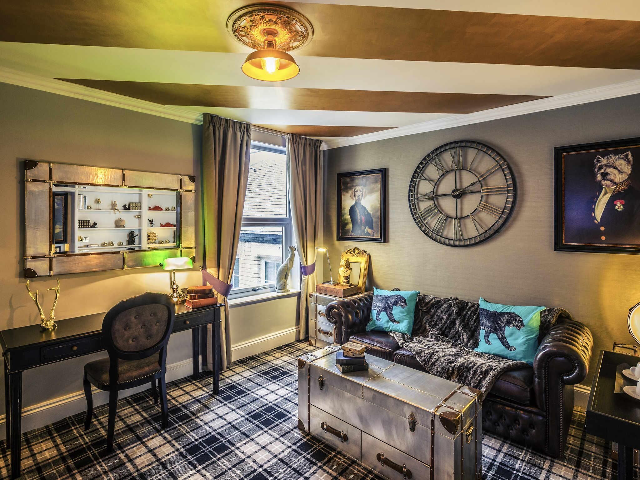 A room with a checkered floor, a leather sofa, a vintage suitcase, a desk with a lamp, and a wall clock.