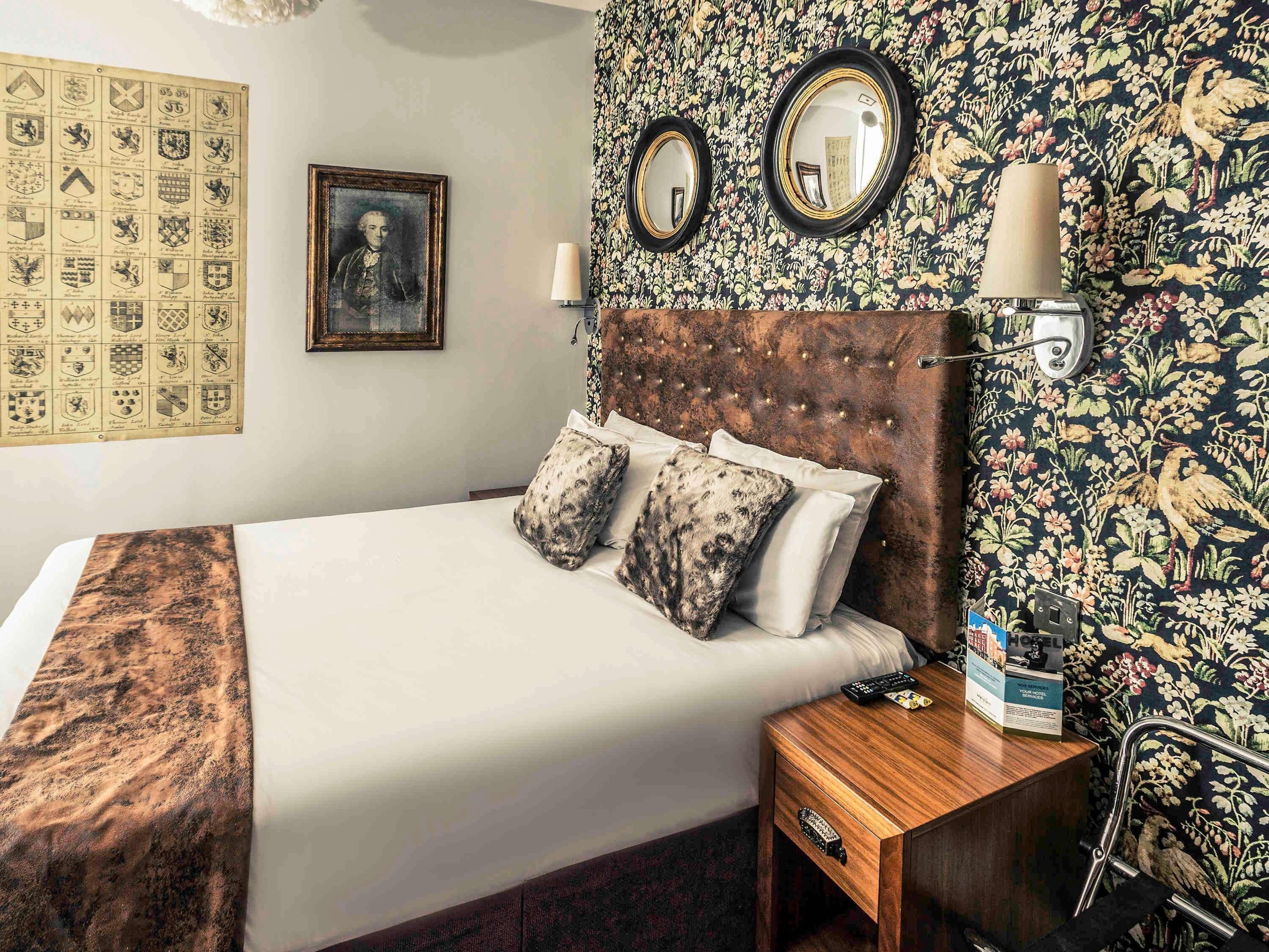 Una habitación de hotel con una cama grande, una mesita de noche de madera, un espejo y un papel tapiz con motivos florales.
