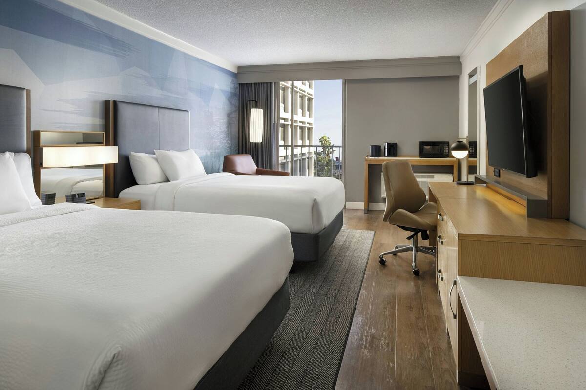 A hotel room with two beds, a desk, a TV, and a view of the city.