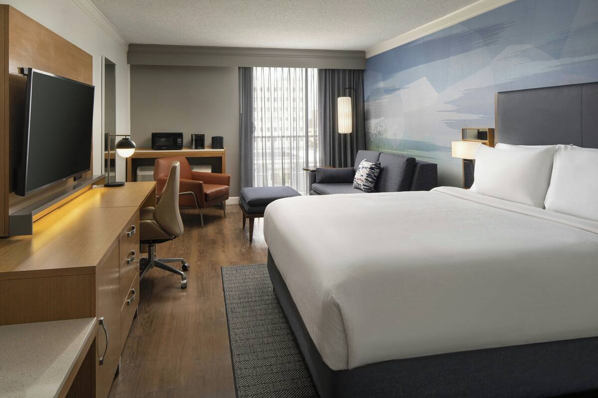 A hotel room with a large bed, a desk with a flat-screen TV, a comfortable armchair, and a view of the city through the window.