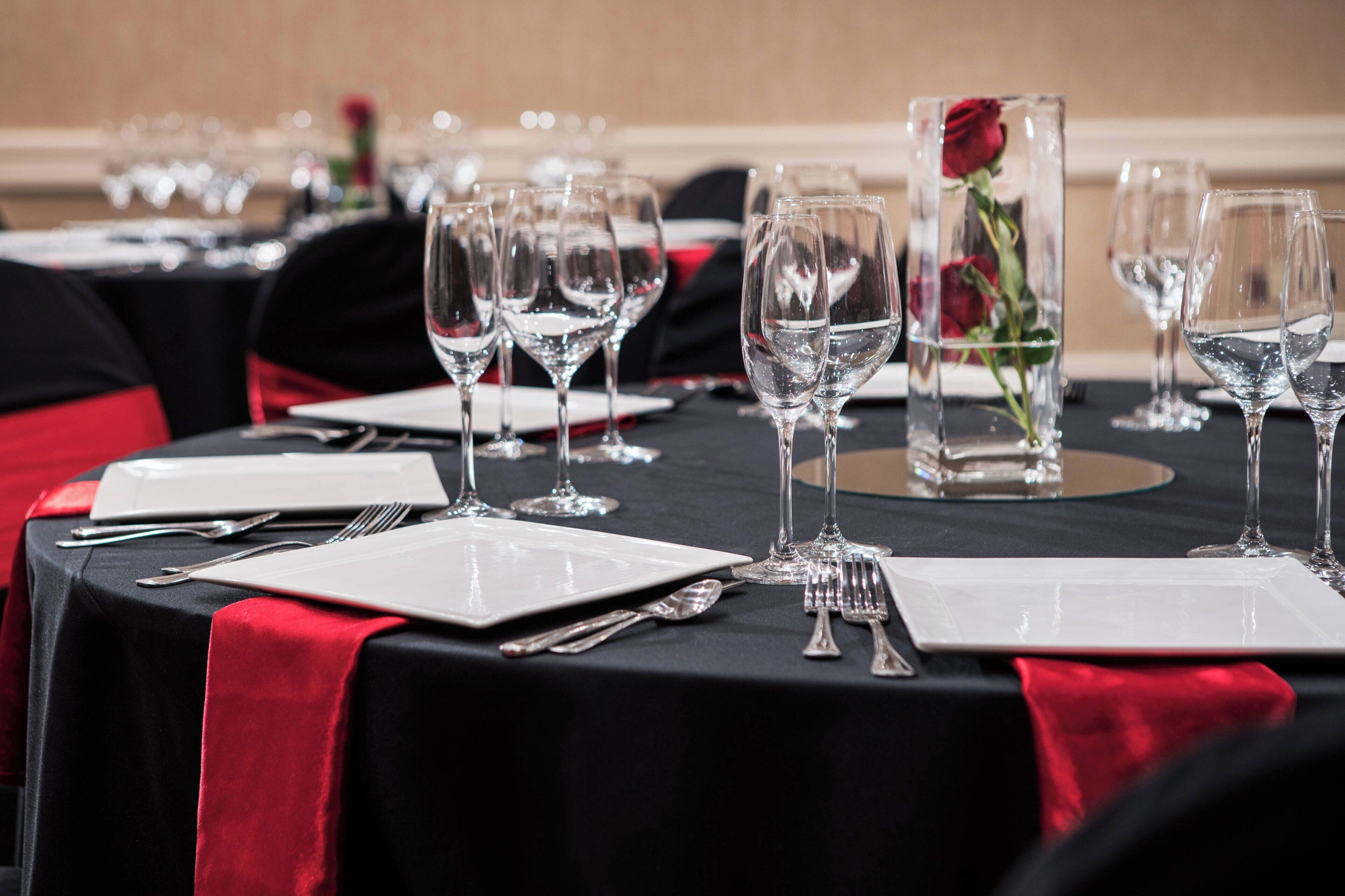 A table set with wine glasses, plates, and cutlery, with a red table runner and a vase with a red rose.