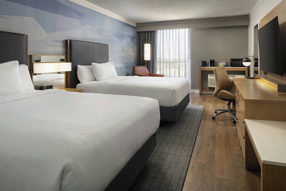 A hotel room with two beds, a desk, a chair, a TV, and a large window with curtains.
