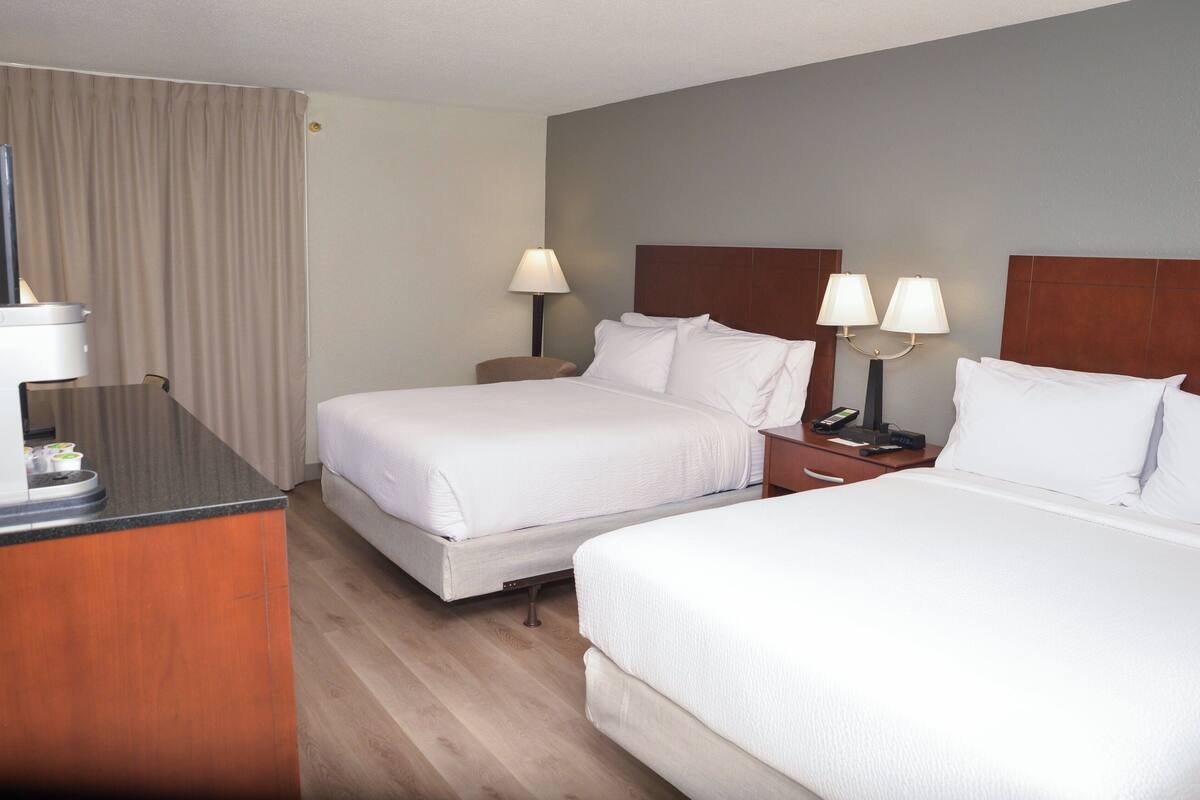 Une chambre d’hôtel avec deux lits, un bureau et une chaise. Il y a deux tables de chevet avec des lampes.