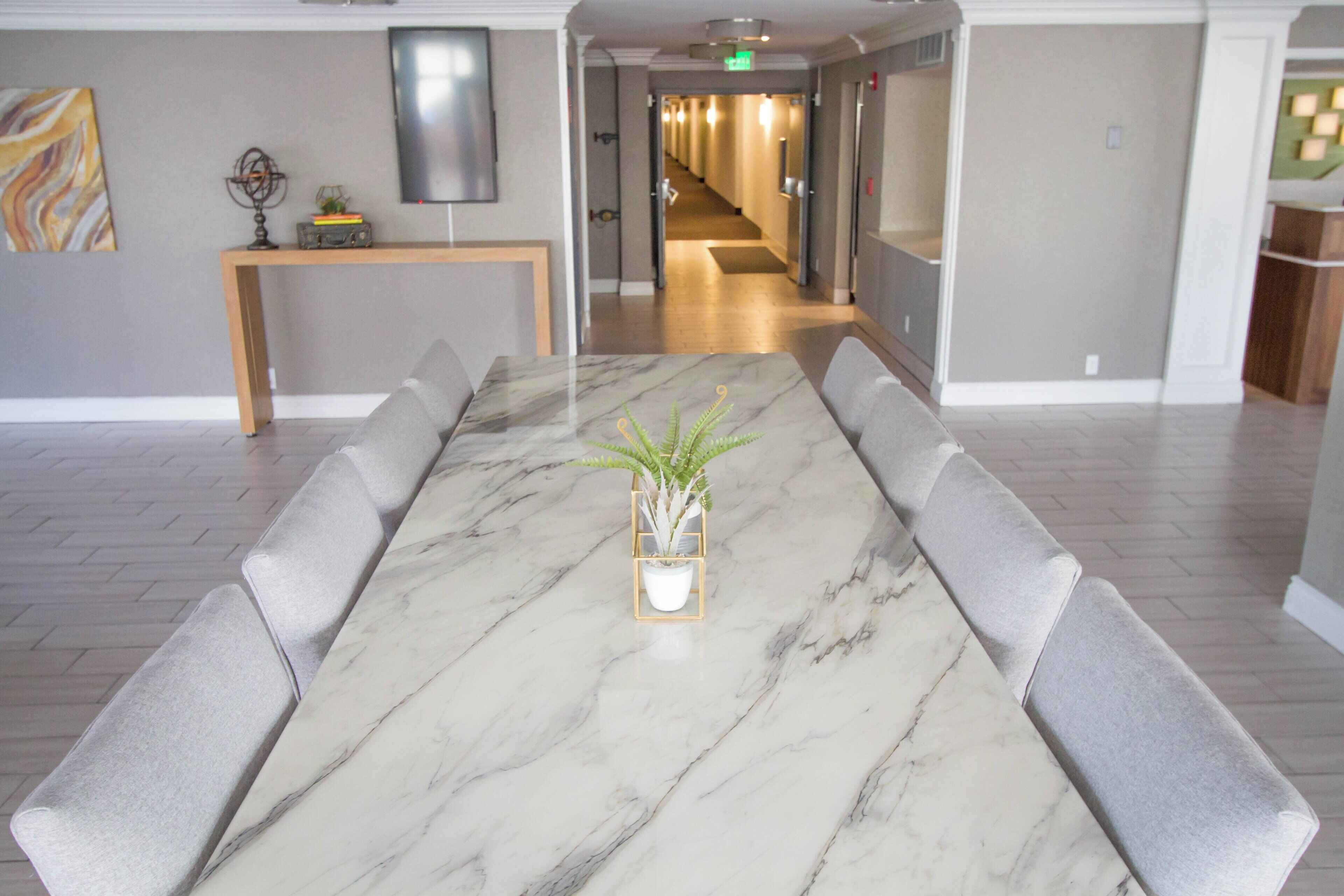 A conference room with a marble table, grey cushioned chairs, a potted plant, and a TV on the wall.