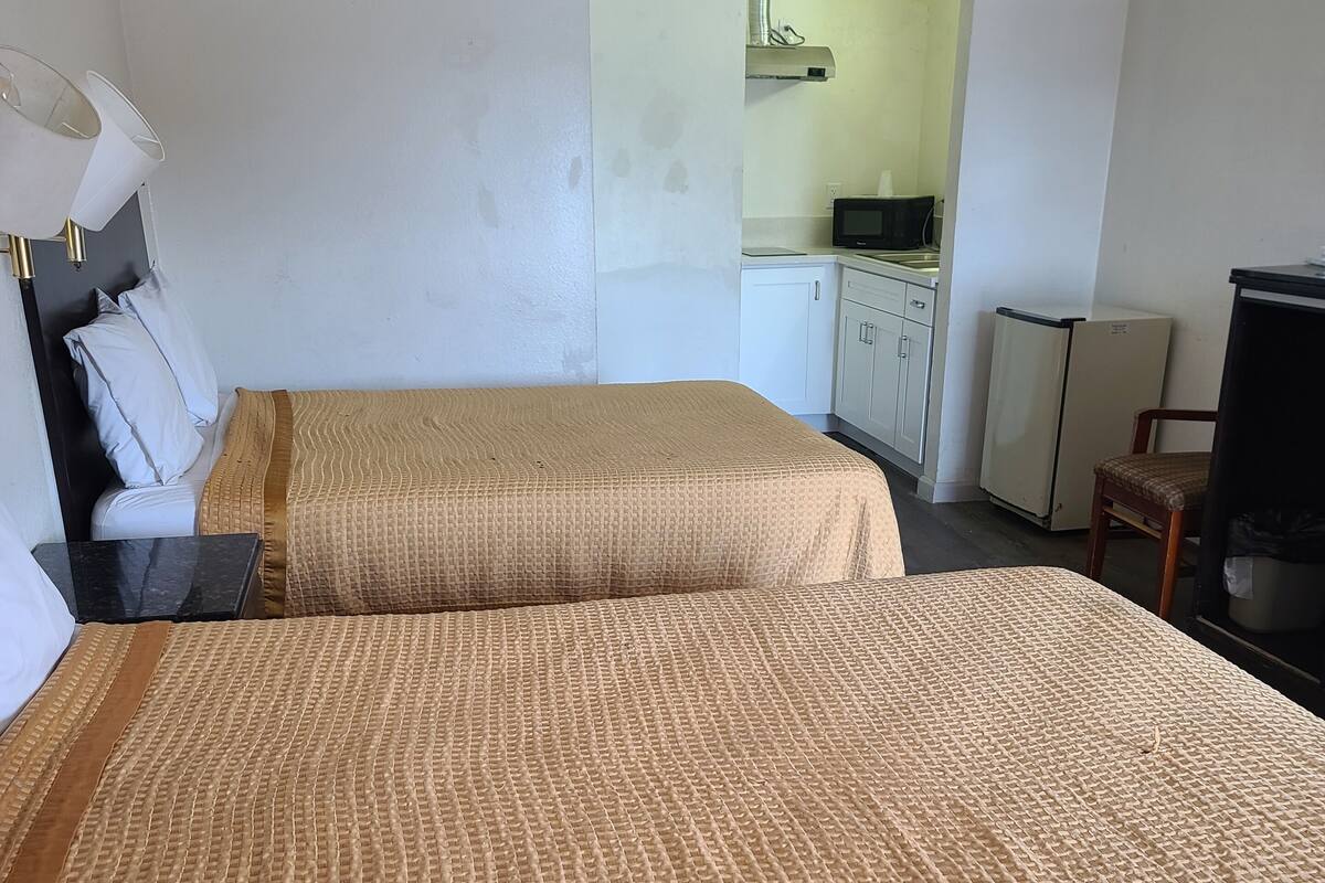 A room with two beds, a desk, a chair, a television, and a kitchenette in the background.