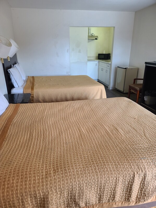 A room with two beds, a desk, a chair, a television, and a kitchenette in the background.