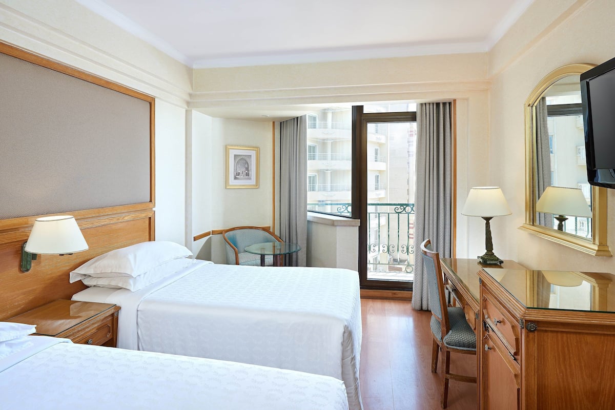 A hotel room with two beds, a desk with a chair, a TV, and a balcony with a view of buildings.