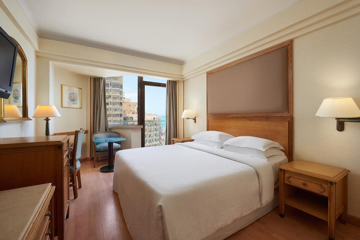 A hotel room with a large bed, a desk with a chair, a TV, and a window with a view of buildings.