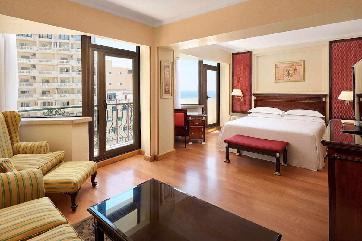A hotel room with a bed, a desk, a chair, a bench, and a balcony with a view of a building.