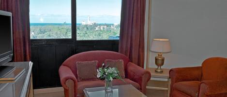 Una habitación con sofá, sillón y una mesa de centro con un jarrón. Un televisor está sobre un mueble bajo, y hay una ventana con cortinas.