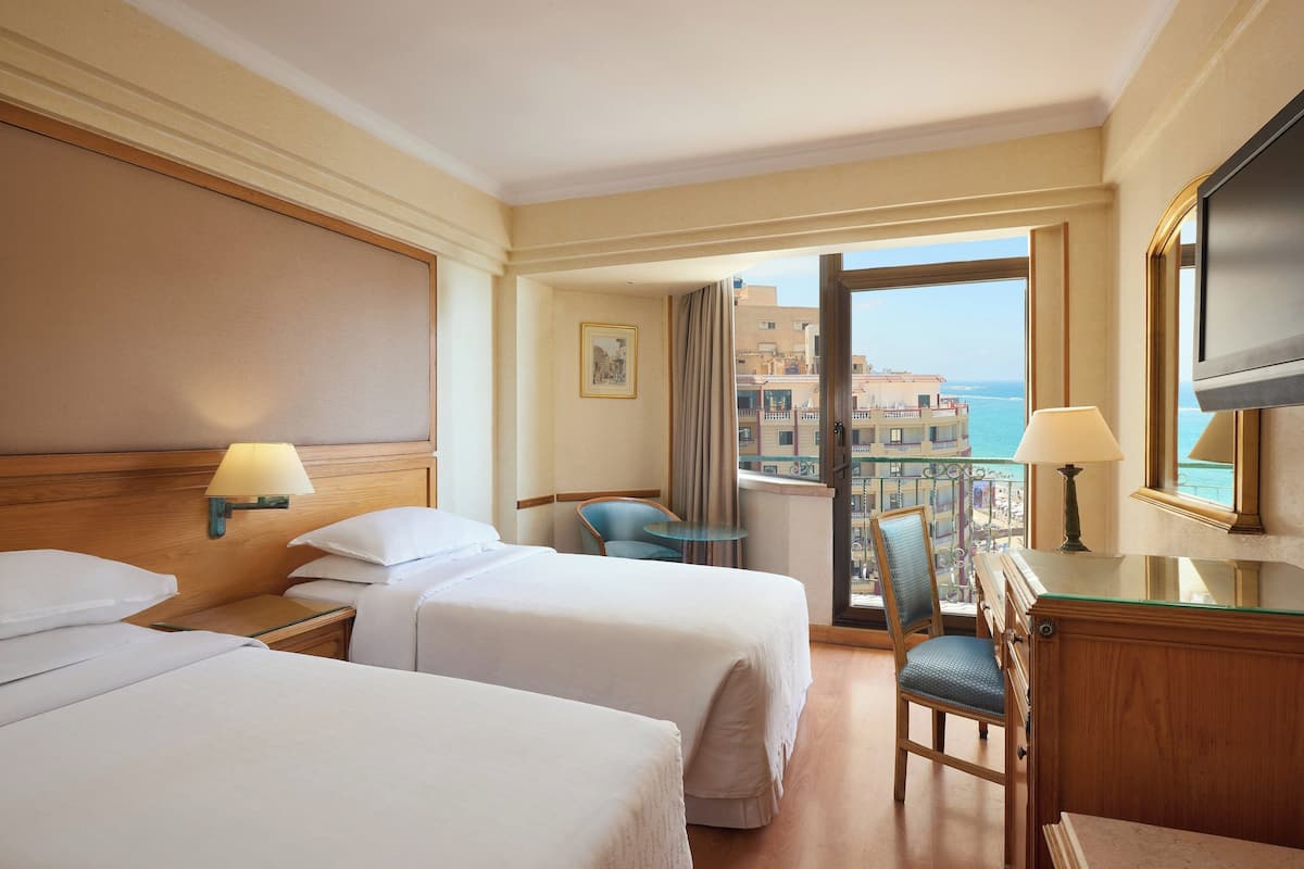 A hotel room with two beds, a desk, a chair, a TV, and a balcony with a view of the sea and buildings.