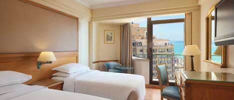 A hotel room with two beds, a desk, a chair, a TV, and a balcony with a view of the sea and buildings.