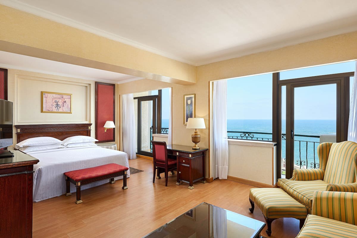 A hotel room with a bed, desk, chair, and a view of the sea.