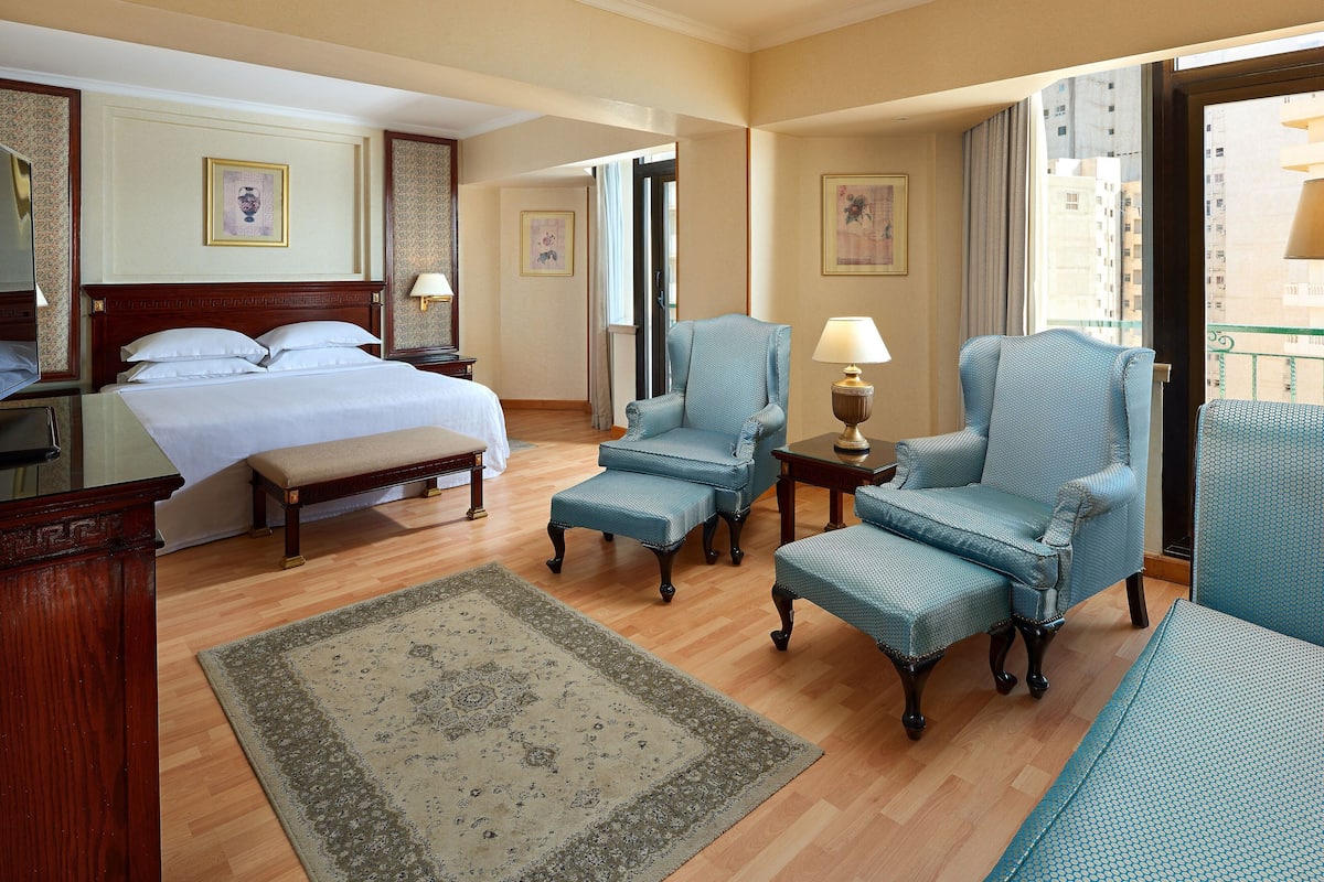 A hotel room with a bed, two armchairs, a bench, a small table with a lamp, and a large window.