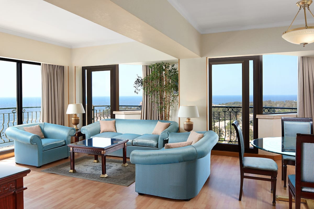 A spacious living room with a blue sofa set, a glass coffee table, and a dining table with chairs. Large windows offer a view of the sea.